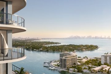 Modern high-rise with curved glass balconies overlooking water and a distant city skyline at sunset.