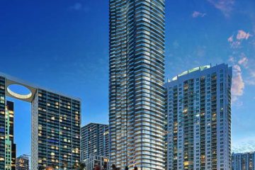 Tall glass skyscraper rises above a marina with luxury yachts under a deep blue evening sky.
