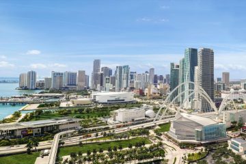 A panoramic view of a coastal city skyline with tall buildings, blue water, and a white arched bridge across the bay.