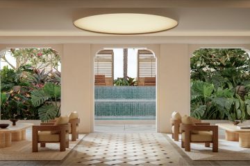 Symmetrical spa lounge with wooden chairs and low tables around beige rugs, tropical plants, and an outdoor water feature visible through arches