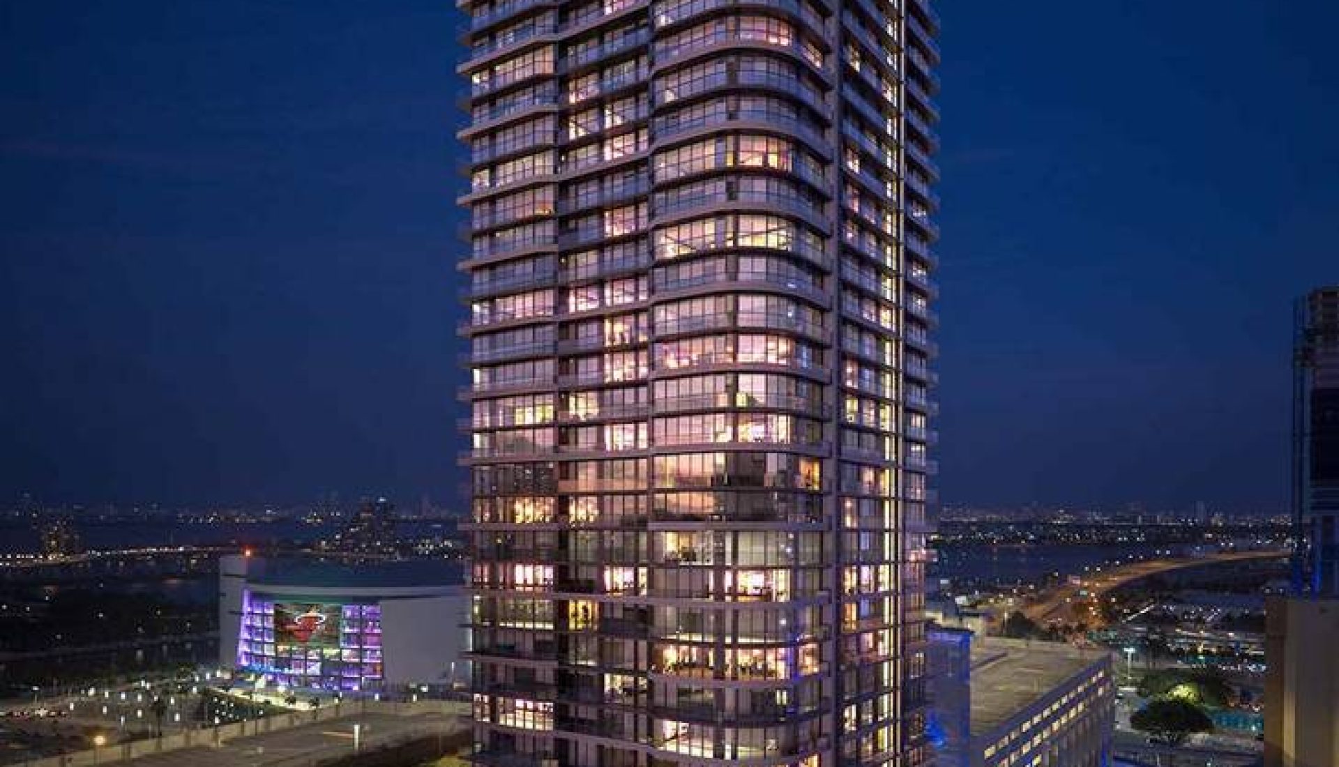 Conceptual rendering of a tall curved glass residential tower at night, illuminated windows, at a busy city intersection.