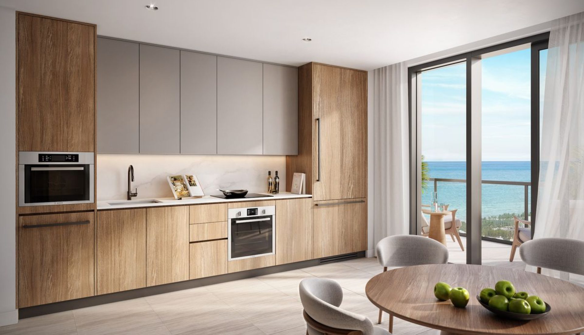 Modern kitchen with wood-toned lower cabinets, gray upper cabinets, and stainless steel appliances; round dining table with apples and a sea-view balcony ahead.