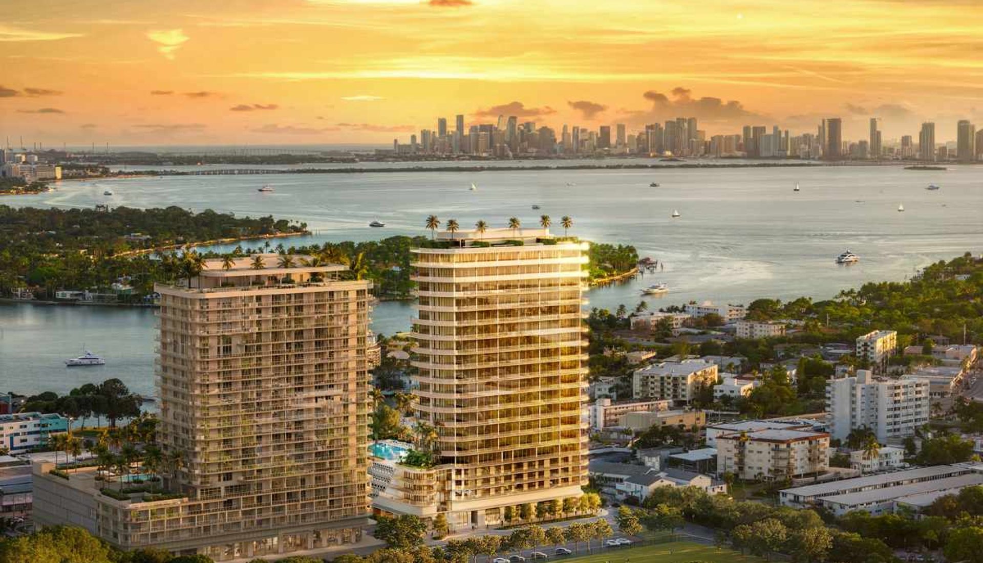 Aerial view of two curved modern high-rise buildings by the water at sunset, with a distant city skyline across the bay.