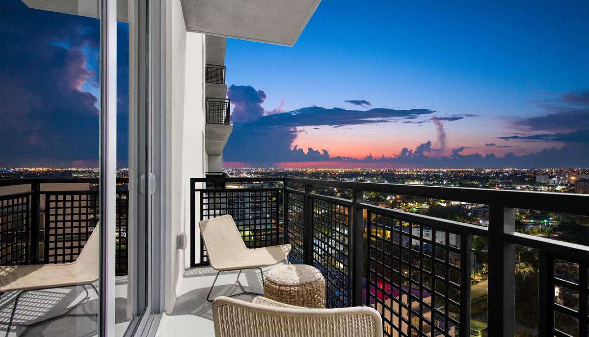 Balcony with two beige chairs and a small wicker table overlooking a city skyline at sunset.