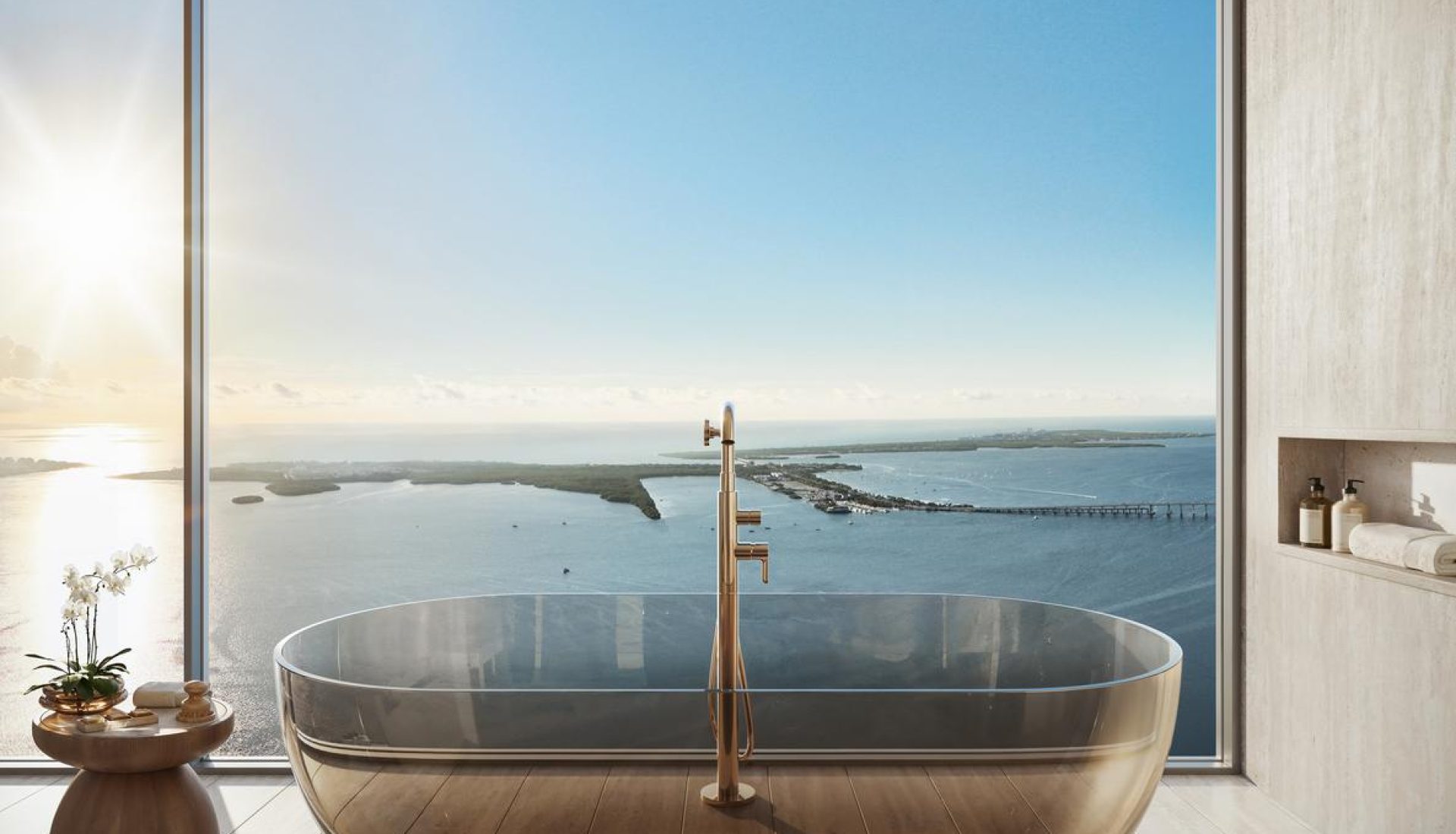 Luxurious bathroom with a glass-walled freestanding tub and brass fixtures, overlooking an ocean coastline at sunrise through a large window.