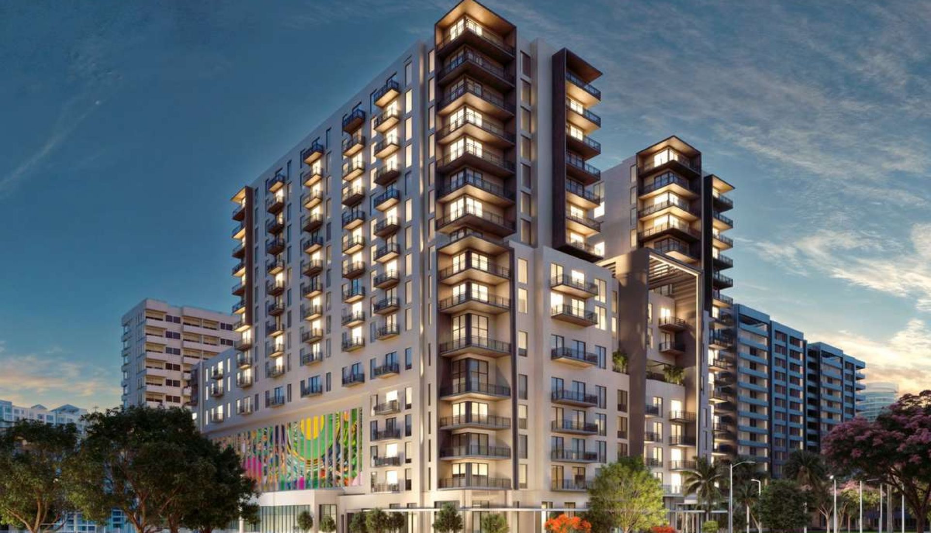 Modern multi-story residential complex with illuminated balconies and a colorful mural on the ground floor at dusk.