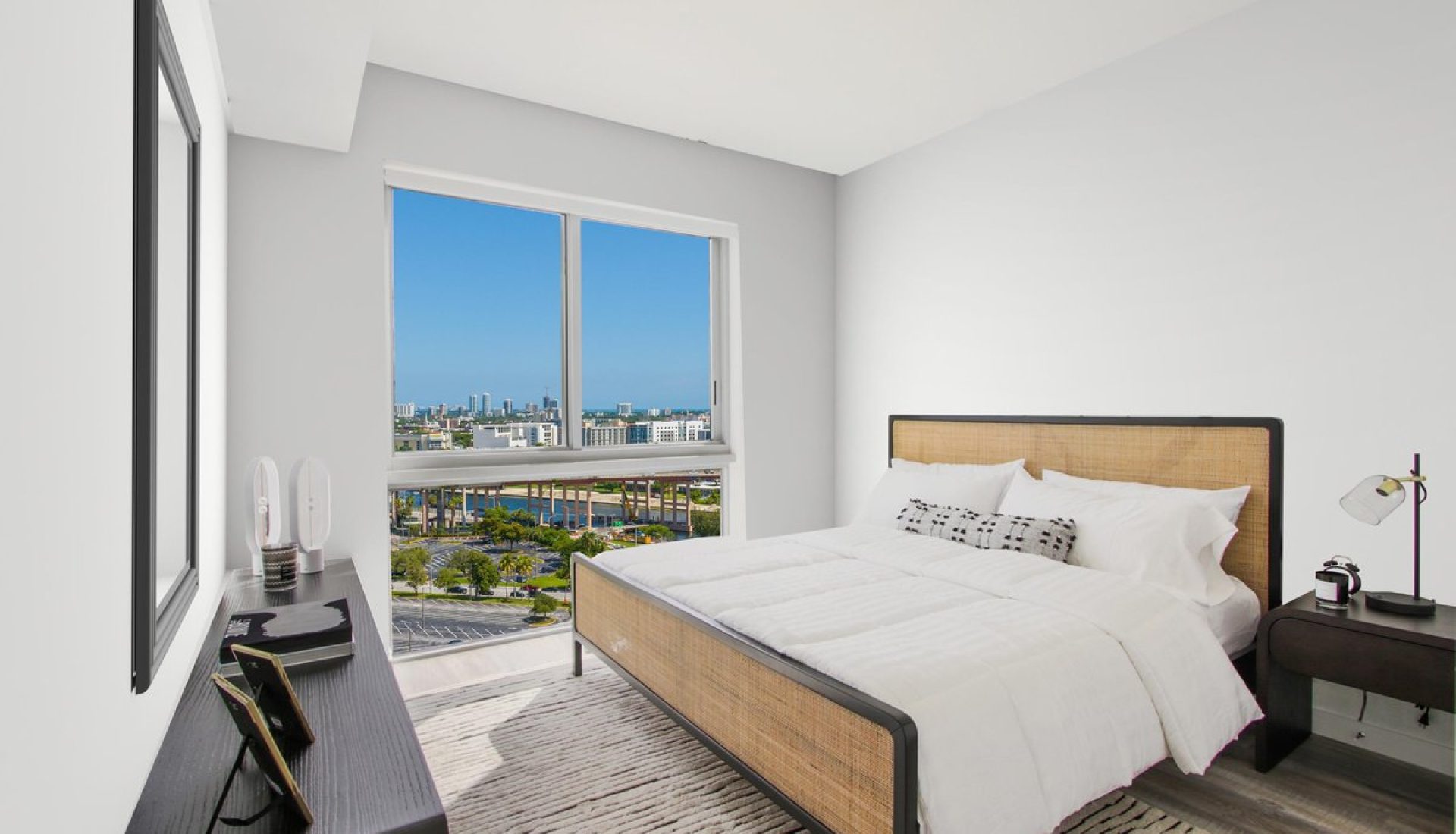 Bright hotel bedroom with a large window offering a city view, white bedding, and a woven-headboard bed�orward.