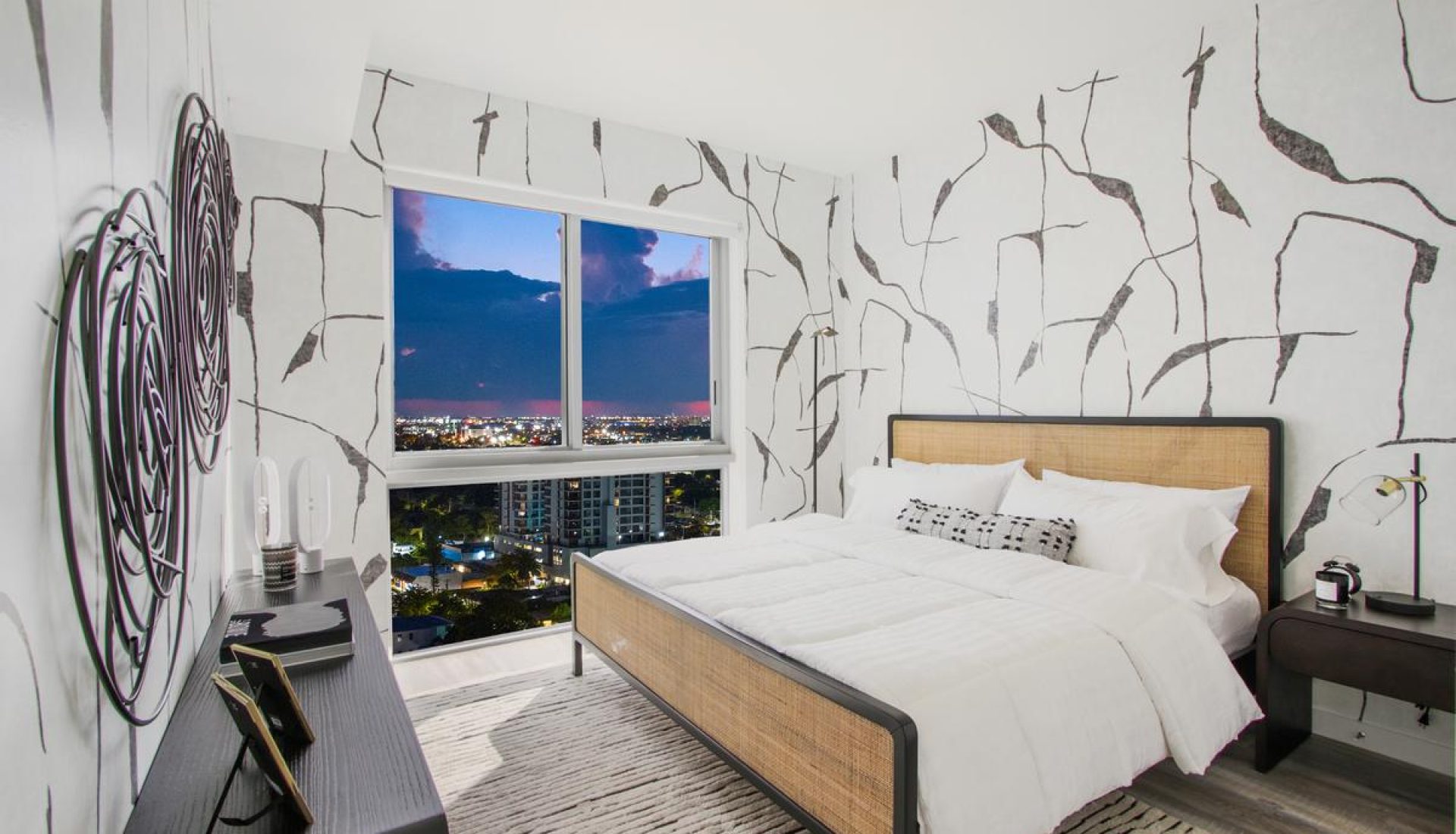 Hotel bedroom with white bedding, woven headboard, and a large window showing a nighttime cityscape. (informative)