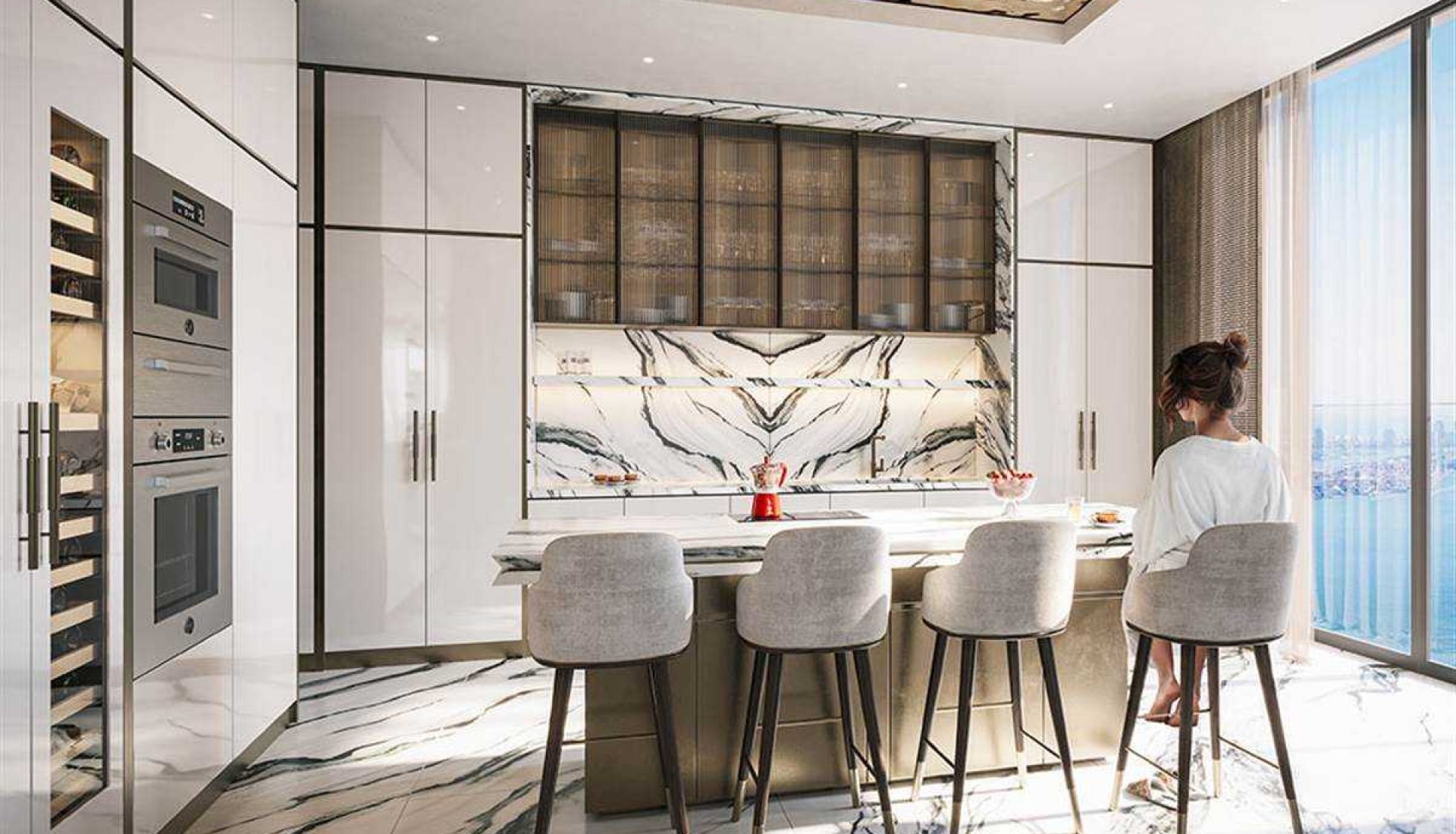 Luxurious modern kitchen with a marble island, high stools, and built-in appliances along white cabinetry; woman sits at the island by a large window overlooking the city.