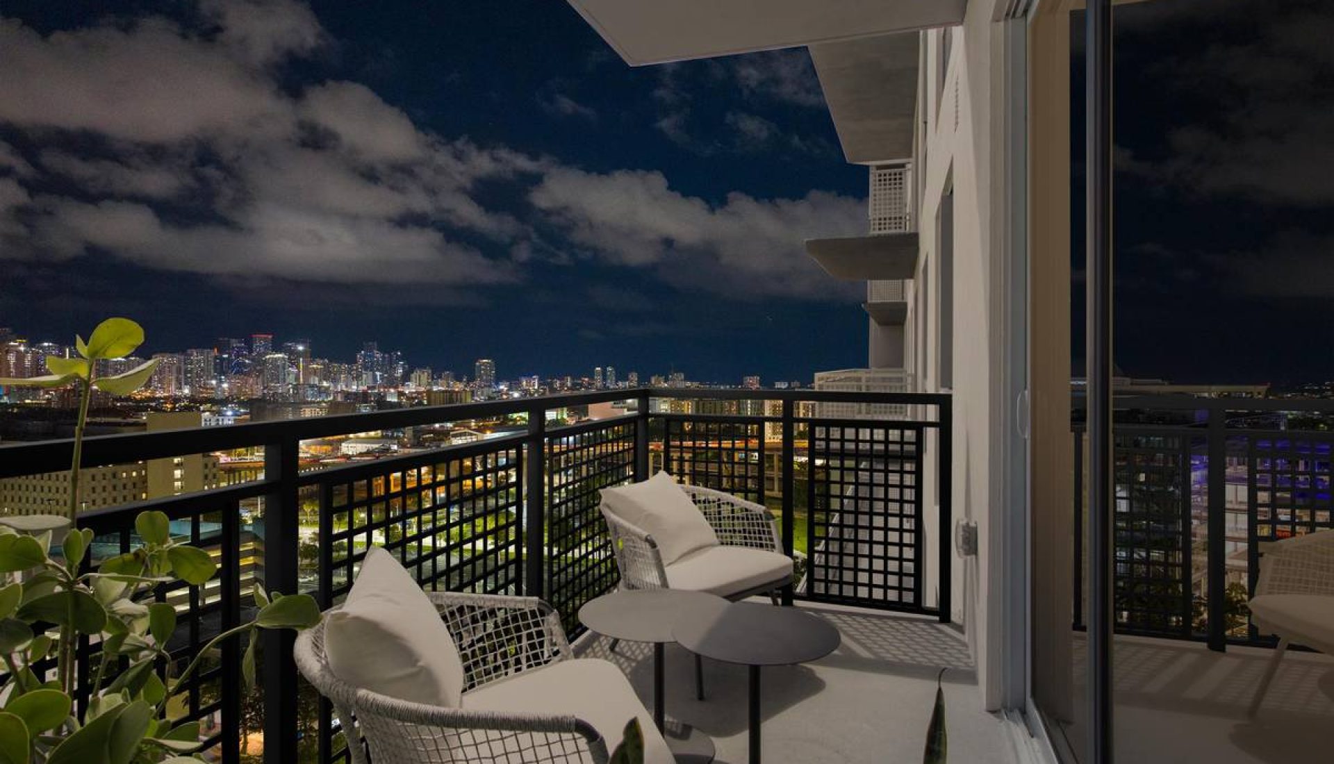 Nighttime city skyline view from a high-rise balcony with white cushioned chairs and a small round table.