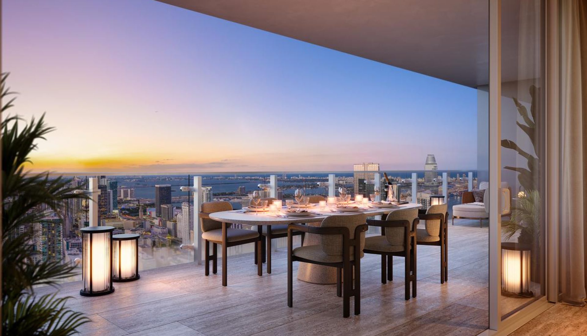 Dinner table set for six on a balcony overlooking a city skyline at sunset, with lit candles on the table.