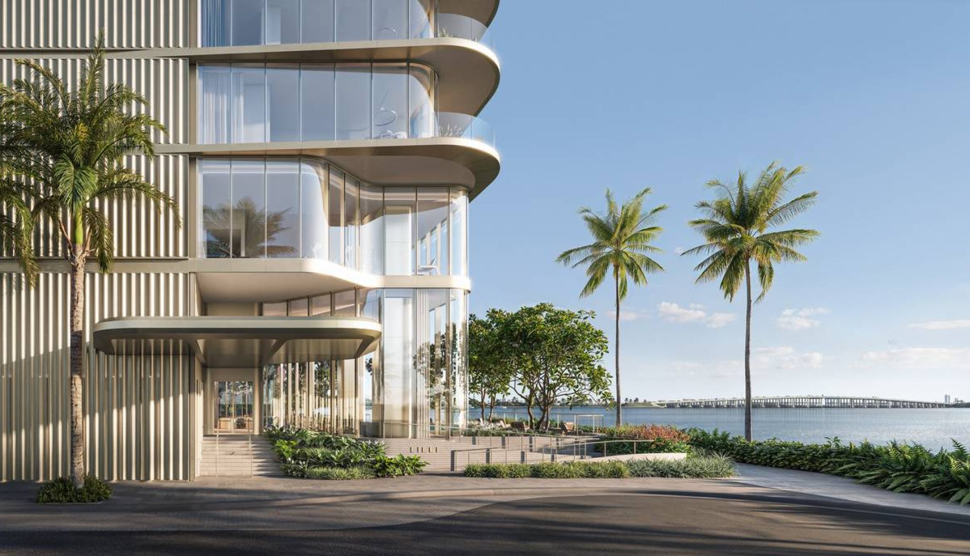 Modern glass-fronted building along a waterfront promenade, with palm trees and landscaped greenery by the water.