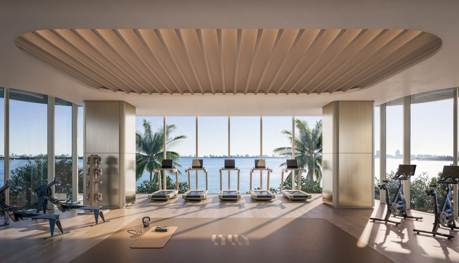 Modern gym with a row of treadmills facing large windows overlooking the ocean and palm trees outside, sunlit space.