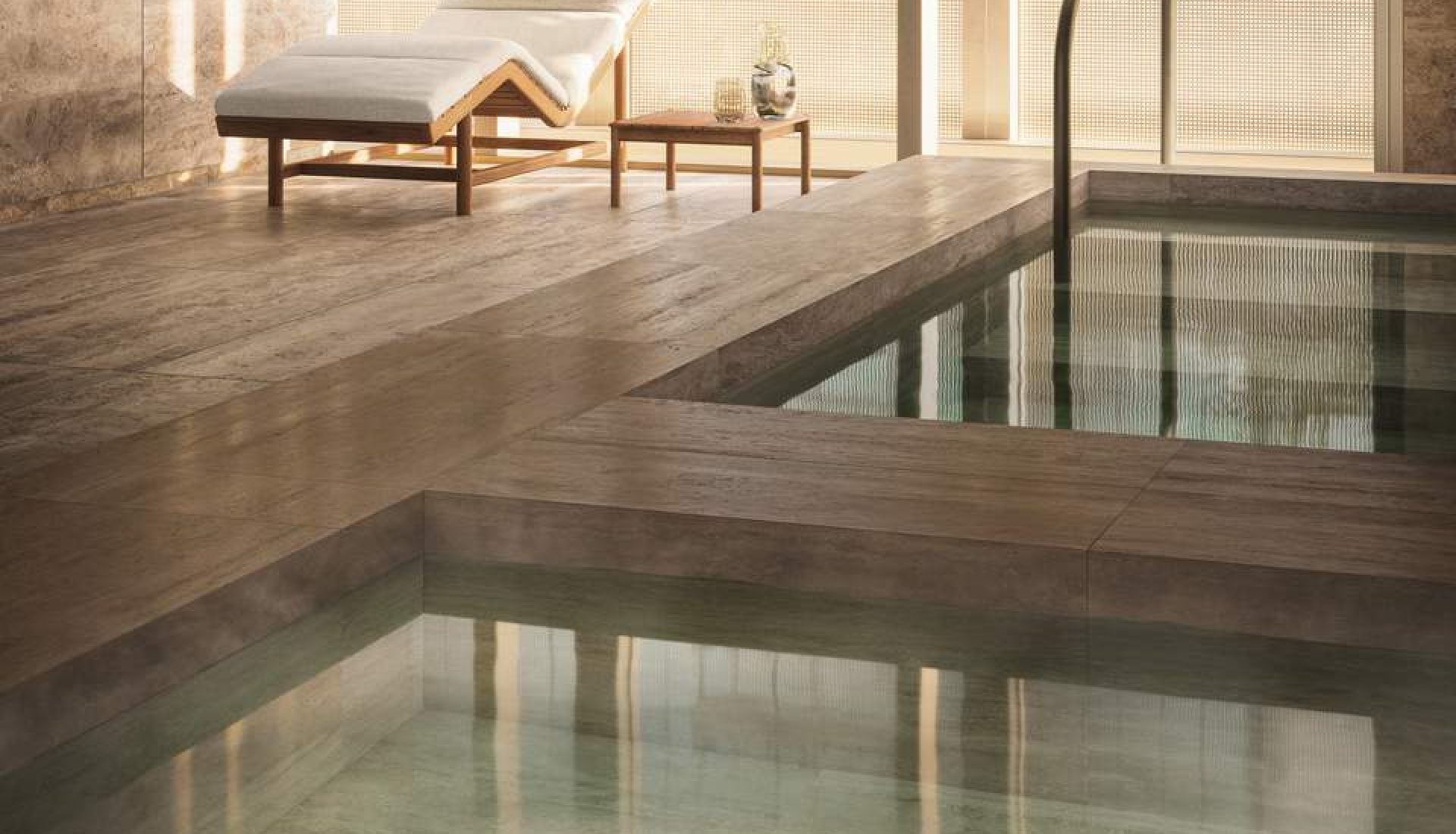Spa lounge area with a wooden chaise, white cushions, and a small table by a warm, sunlit wall. A relaxing indoor pool scene featuring a lounge chair and side table beside a tranquil pool edge.