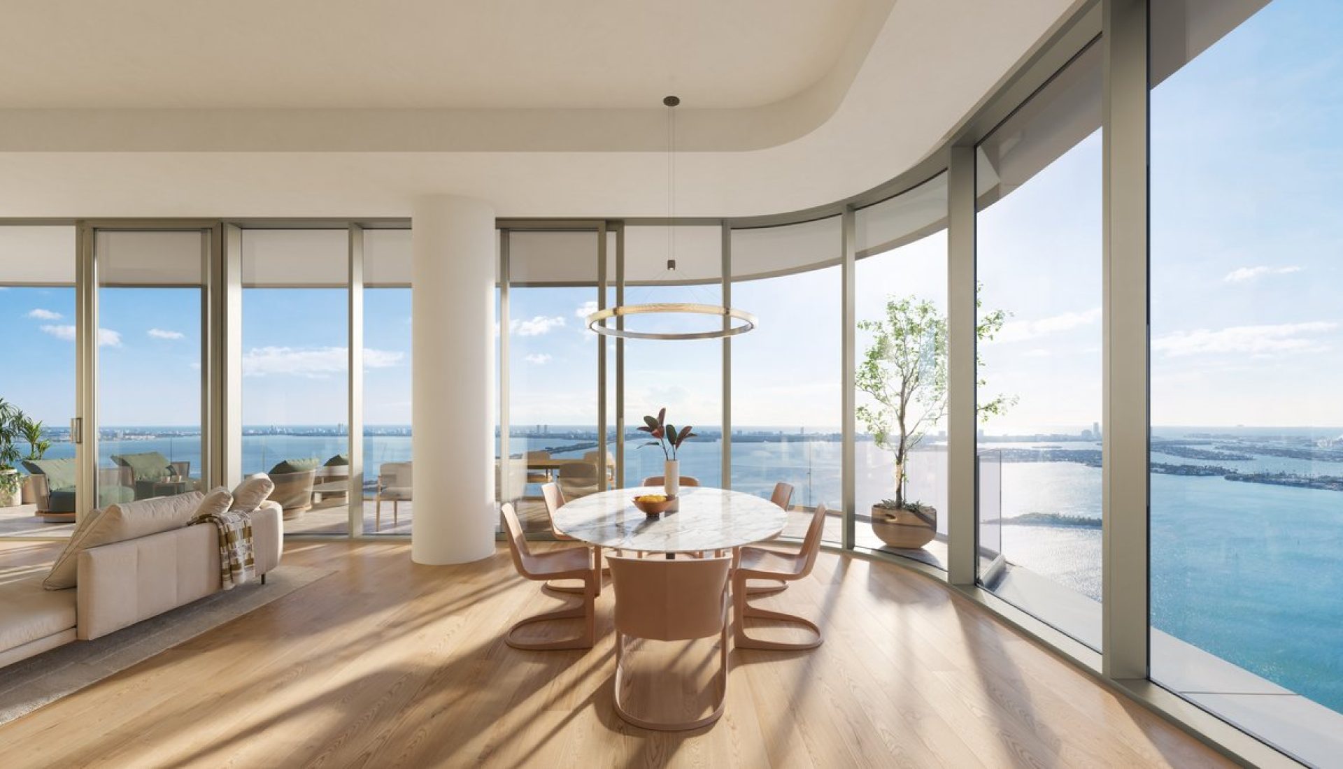 Open-plan living and dining area with a round marble table and pink-beige chairs, curved glass walls, and a panoramic ocean view.