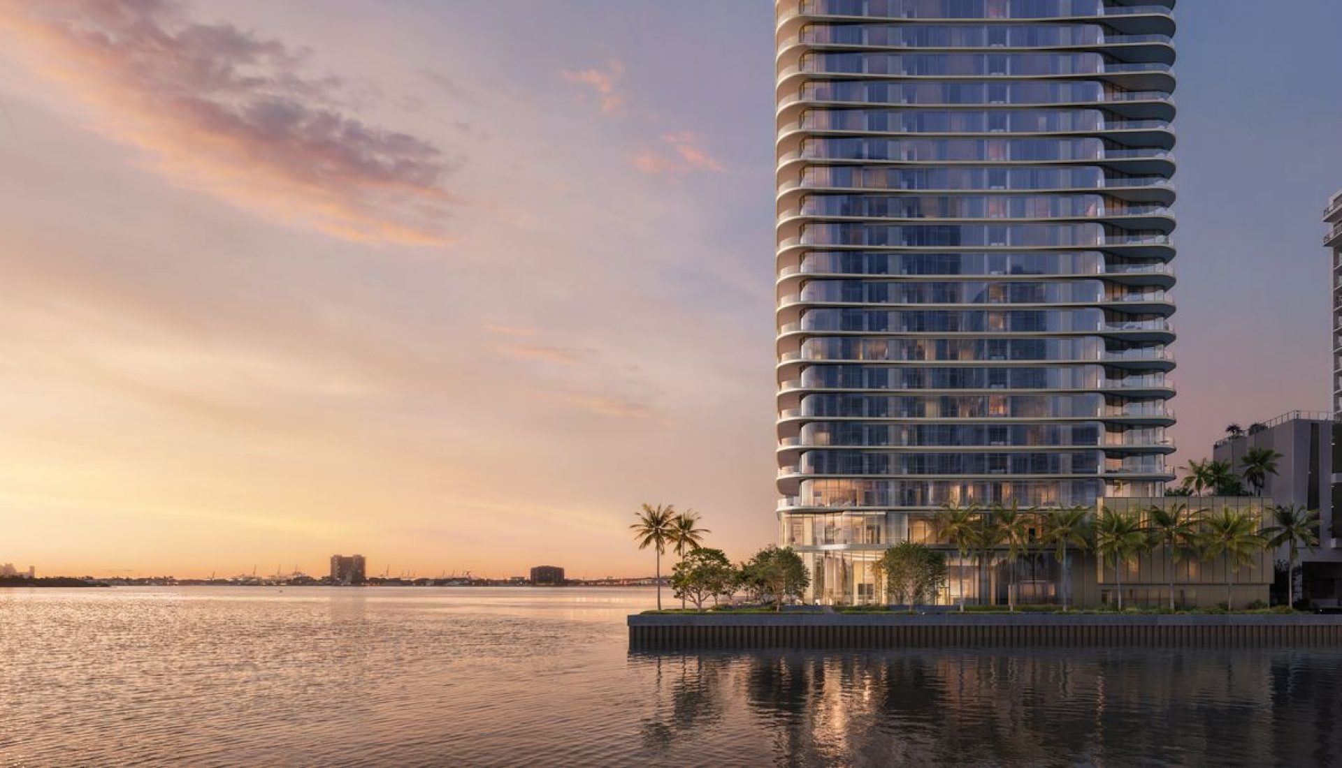 Modern glass high-rise by a calm waterway at sunset, with palm trees along a waterfront promenade.