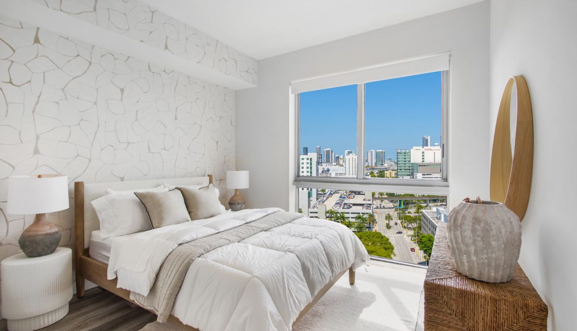 Bright modern bedroom with white bedding and a wooden bed frame, featuring a textured white accent wall and a large window showing a city skyline.