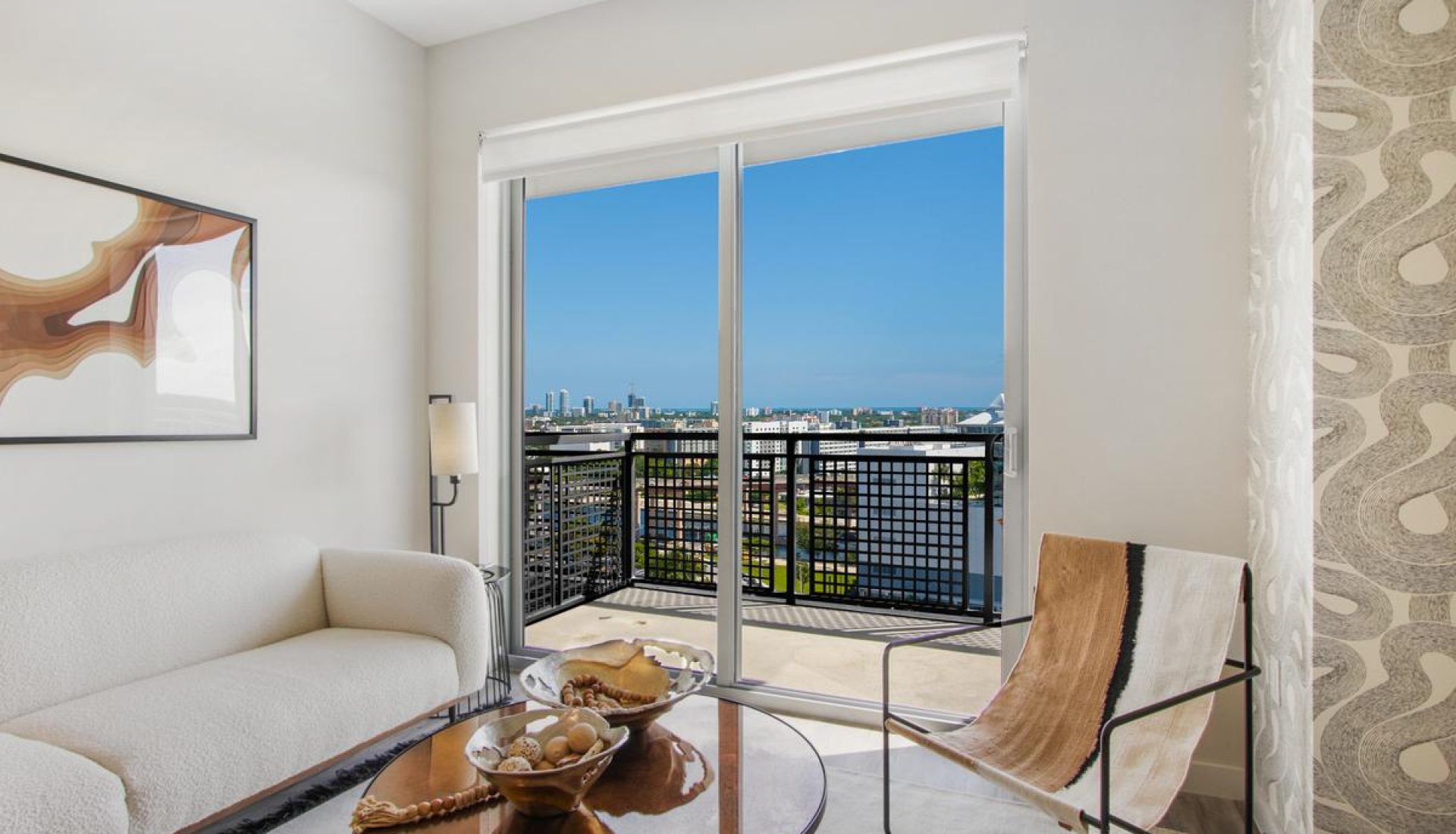 Bright living room with white sofa and glass sliding door opening to a balcony overlooking a city skyline.