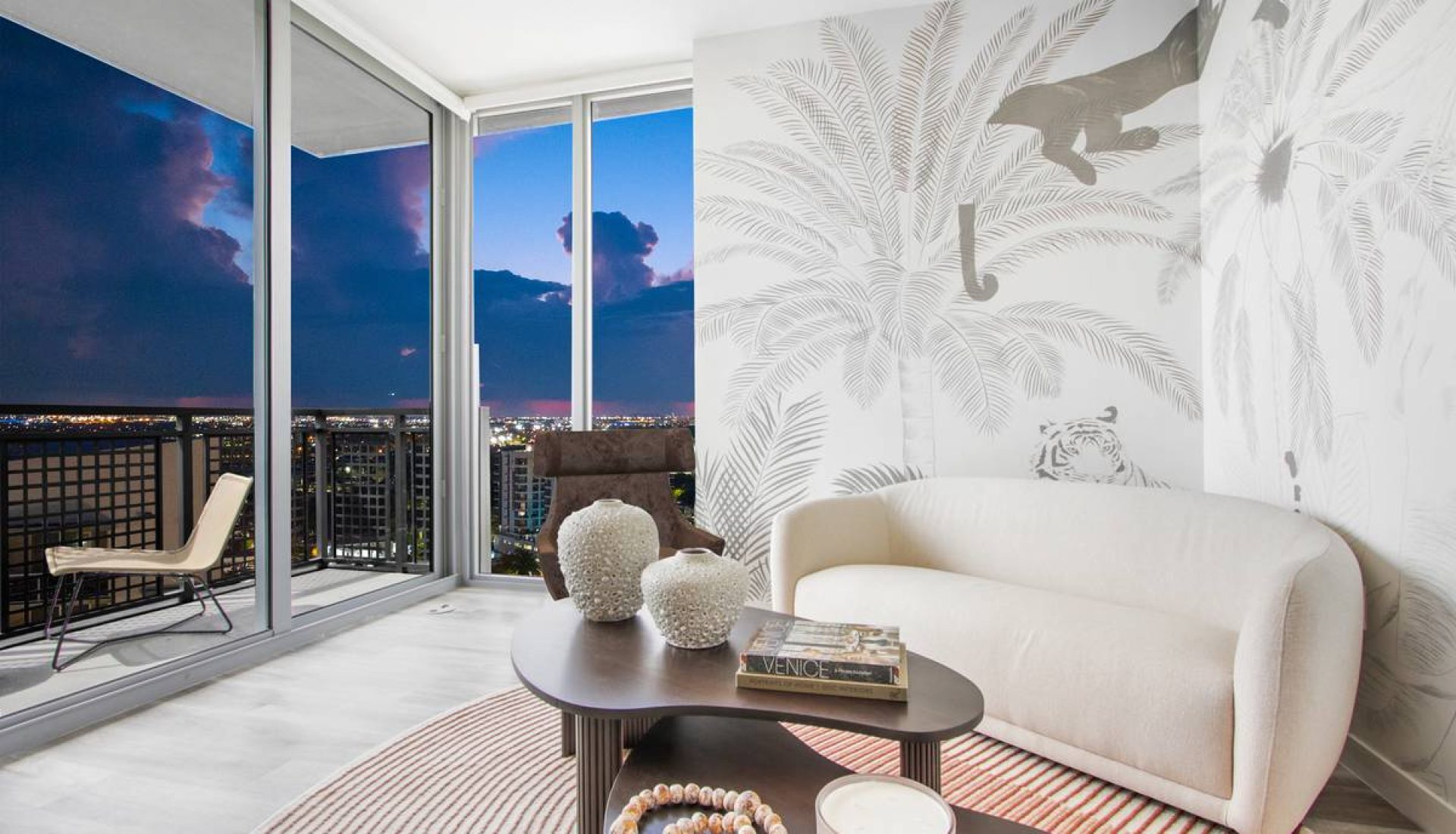 Living room with a white curved sofa, dark wood coffee table, and tropical palm mural; balcony with city lights outside at dusk; decorative vases and a candle on the table.