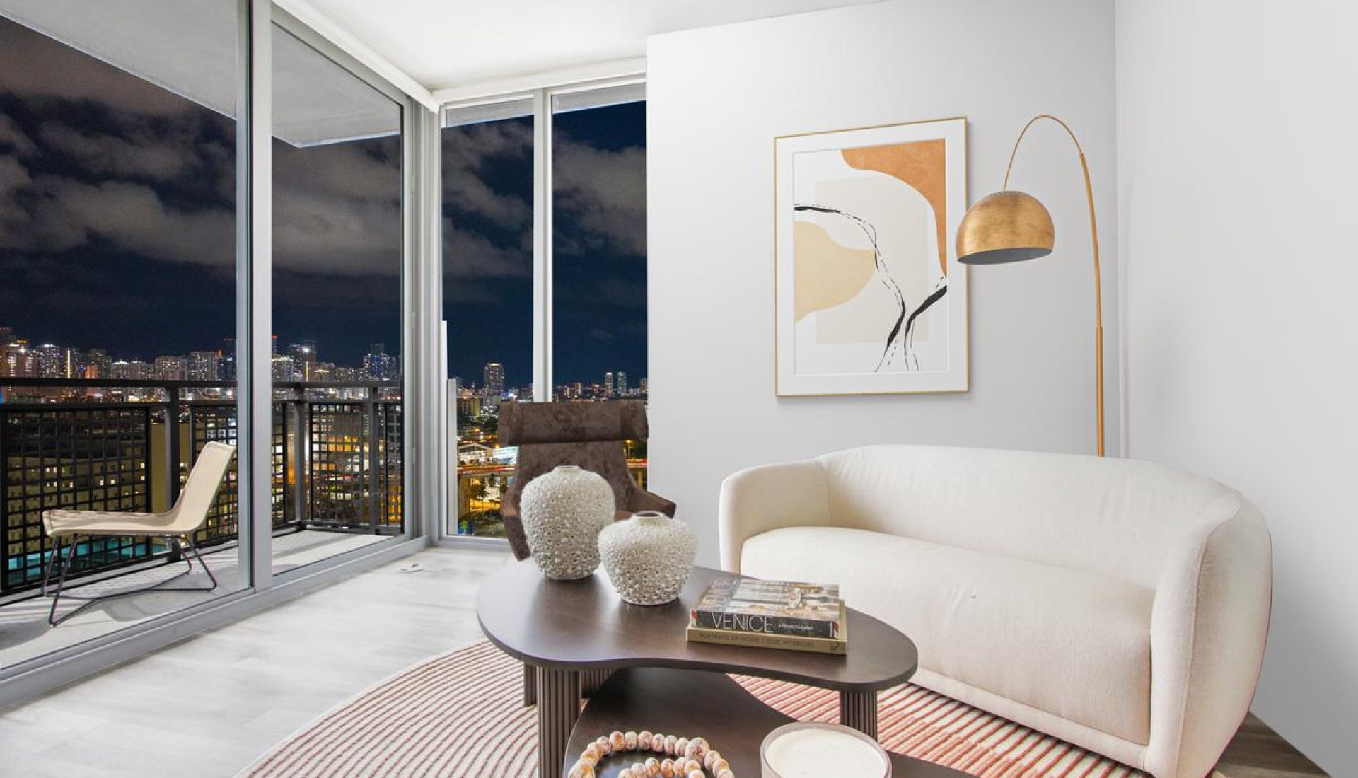 Bright contemporary living room with a cream curved sofa, round coffee table, striped rug, and floor-to-ceiling windows showing a city skyline at night.