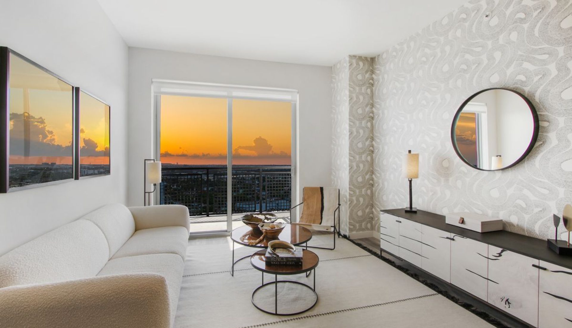 Bright living room with a white sofa, round mirror, and a sunset view through the sliding glass door to the balcony.