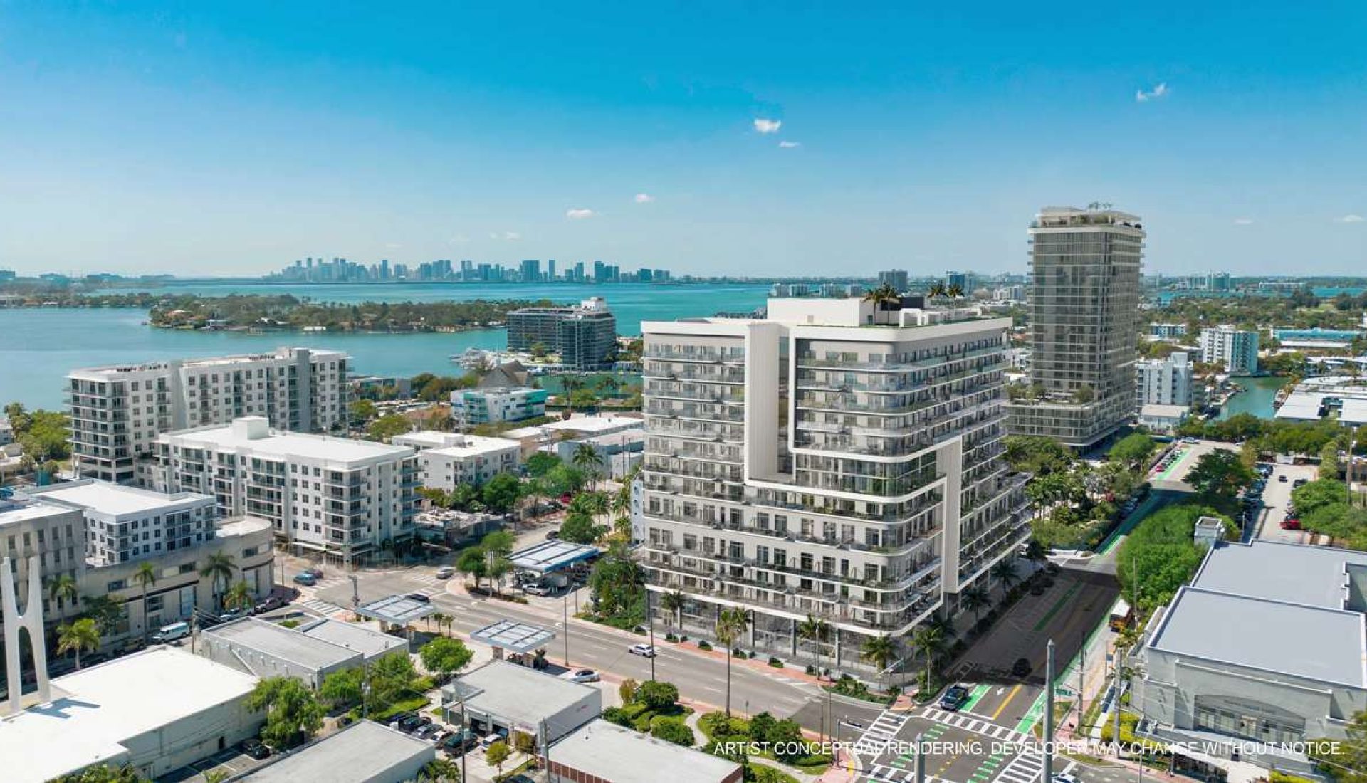 Aerial view of a modern coastal city with a tall white apartment building along a tree-lined street and water in the background.