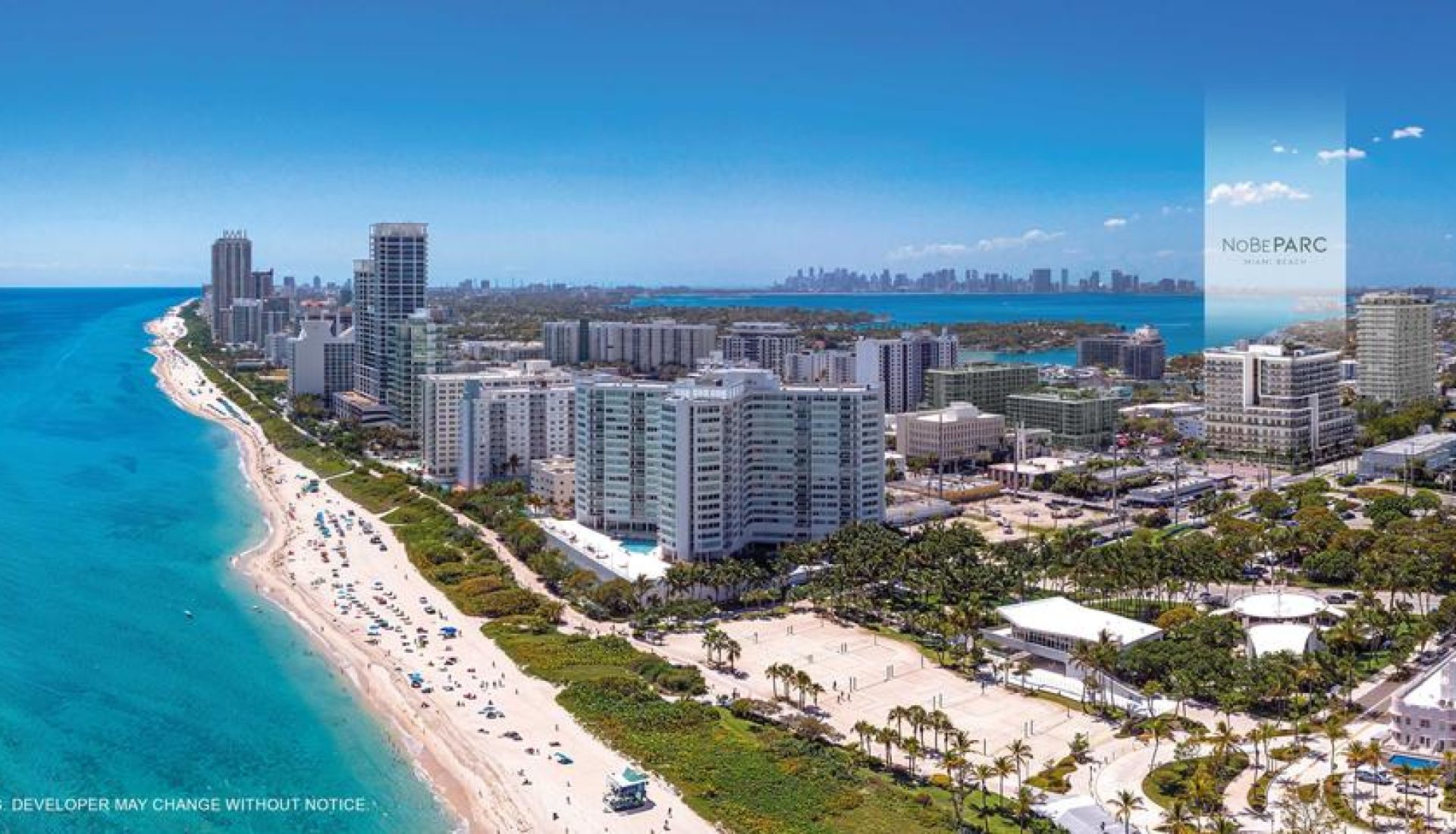 Aerial view of a sunny coastal city with a long sandy beach, turquoise water, and high-rise buildings along the shore.