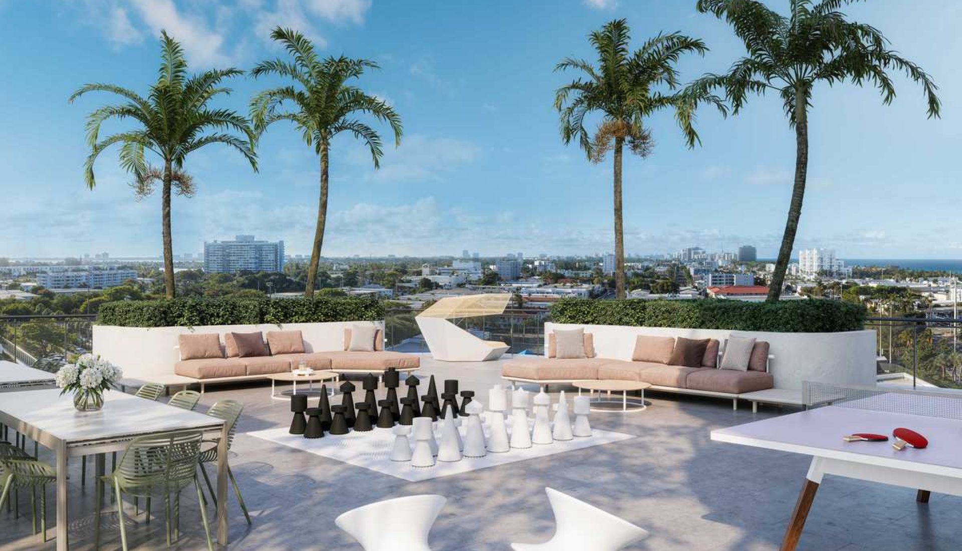 Rooftop lounge with beige sectional sofas, palm trees, and a large outdoor chess set overlooking a city skyline.