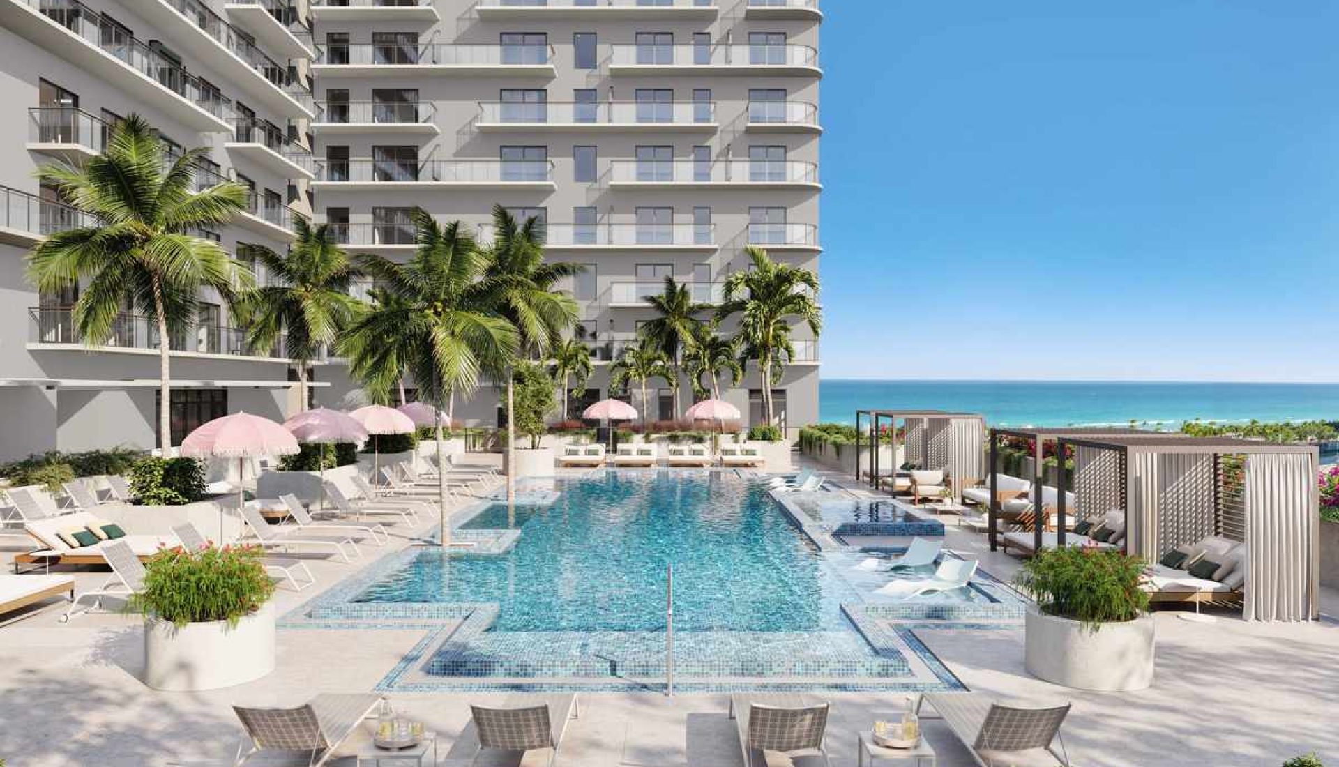 Pool area at a beachfront hotel with palm trees, pink umbrellas, and lounge chairs, and the ocean in the background.