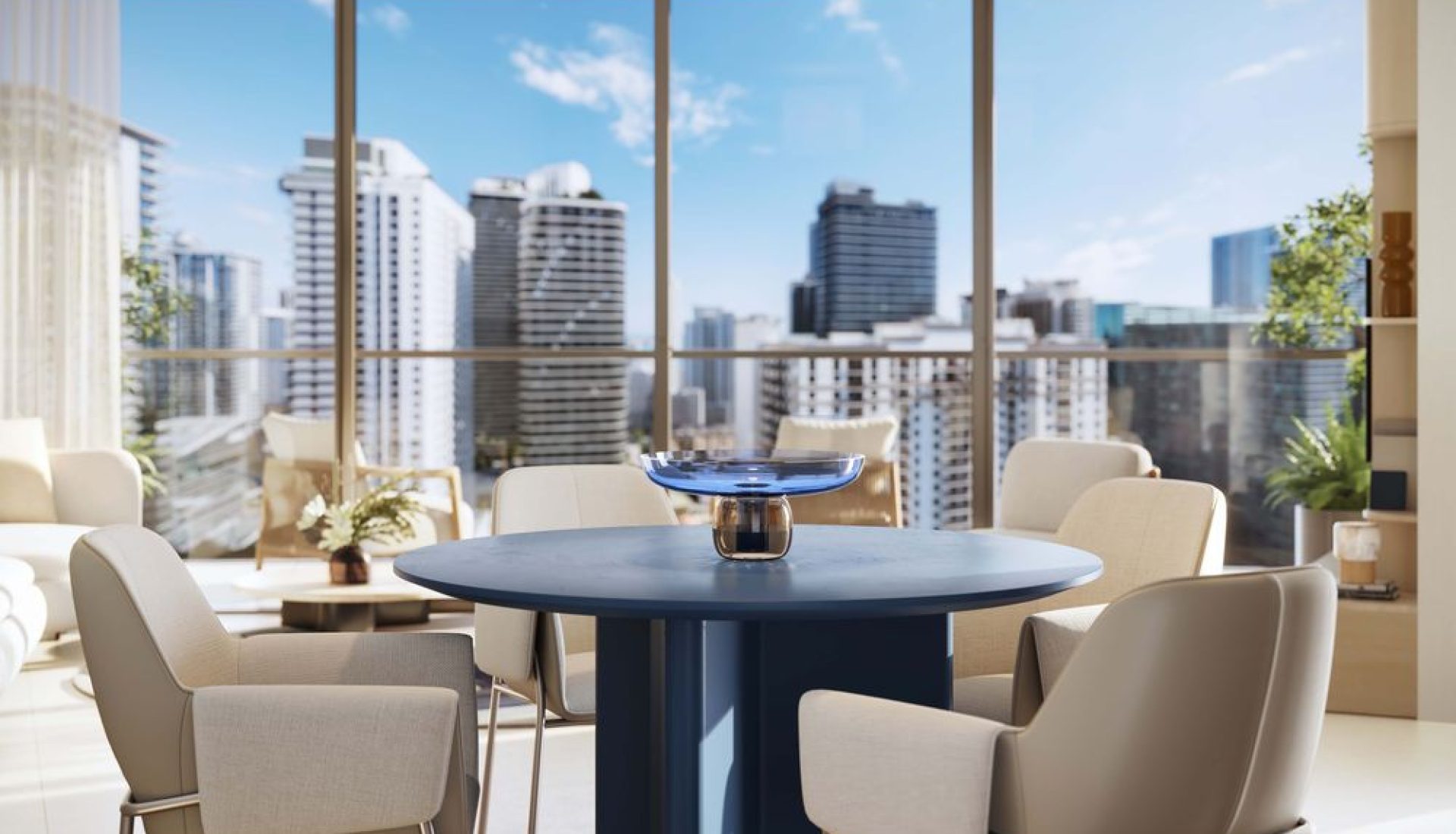 Bright contemporary dining area with a round blue table and six beige upholstered chairs, open to a city skyline through floor‑to‑ceiling windows.