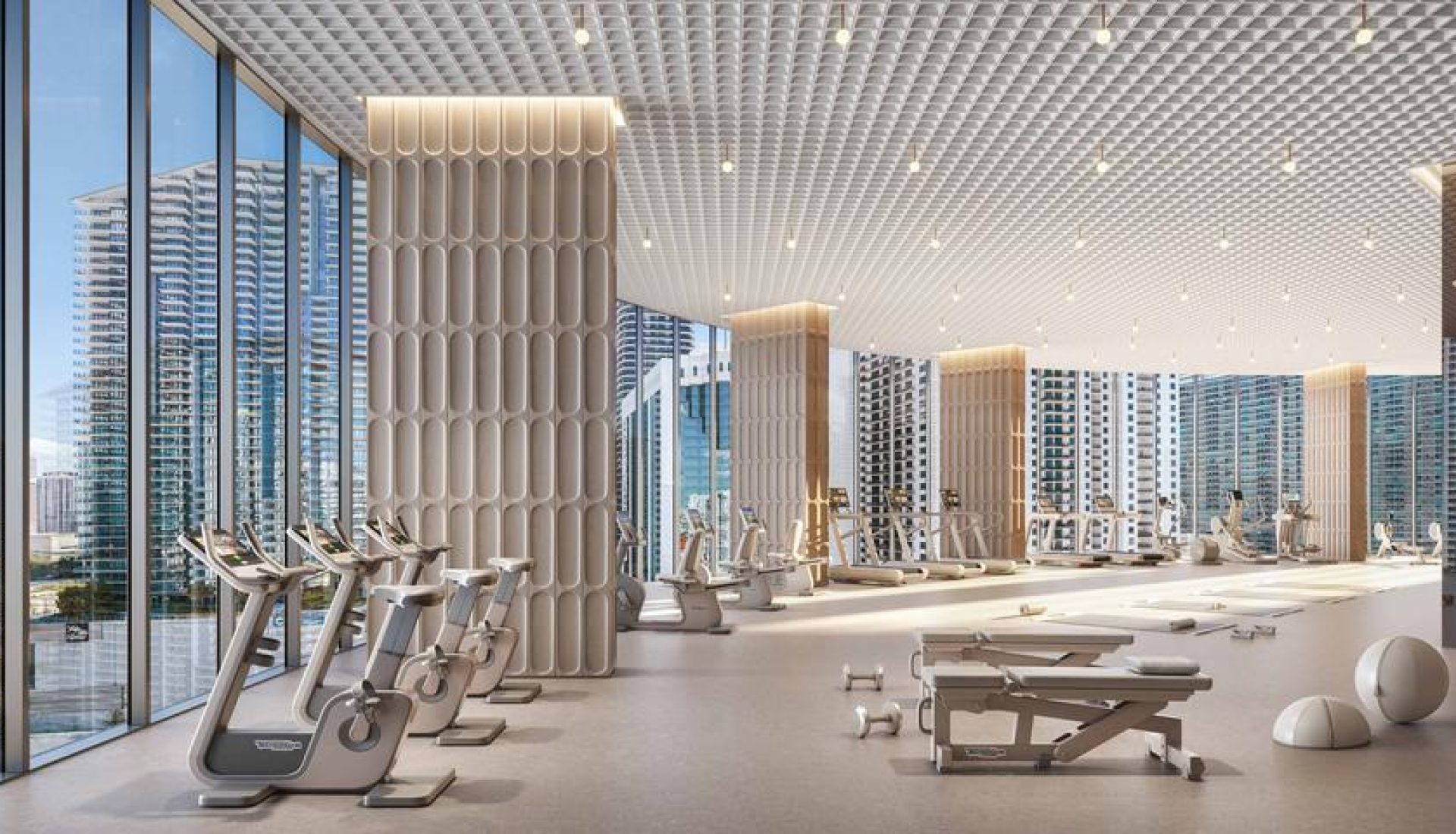 Spacious modern gym with floor-to-ceiling windows overlooking a city waterfront; rows of treadmills and stationary bikes under a grid ceiling, with sculpted columns.