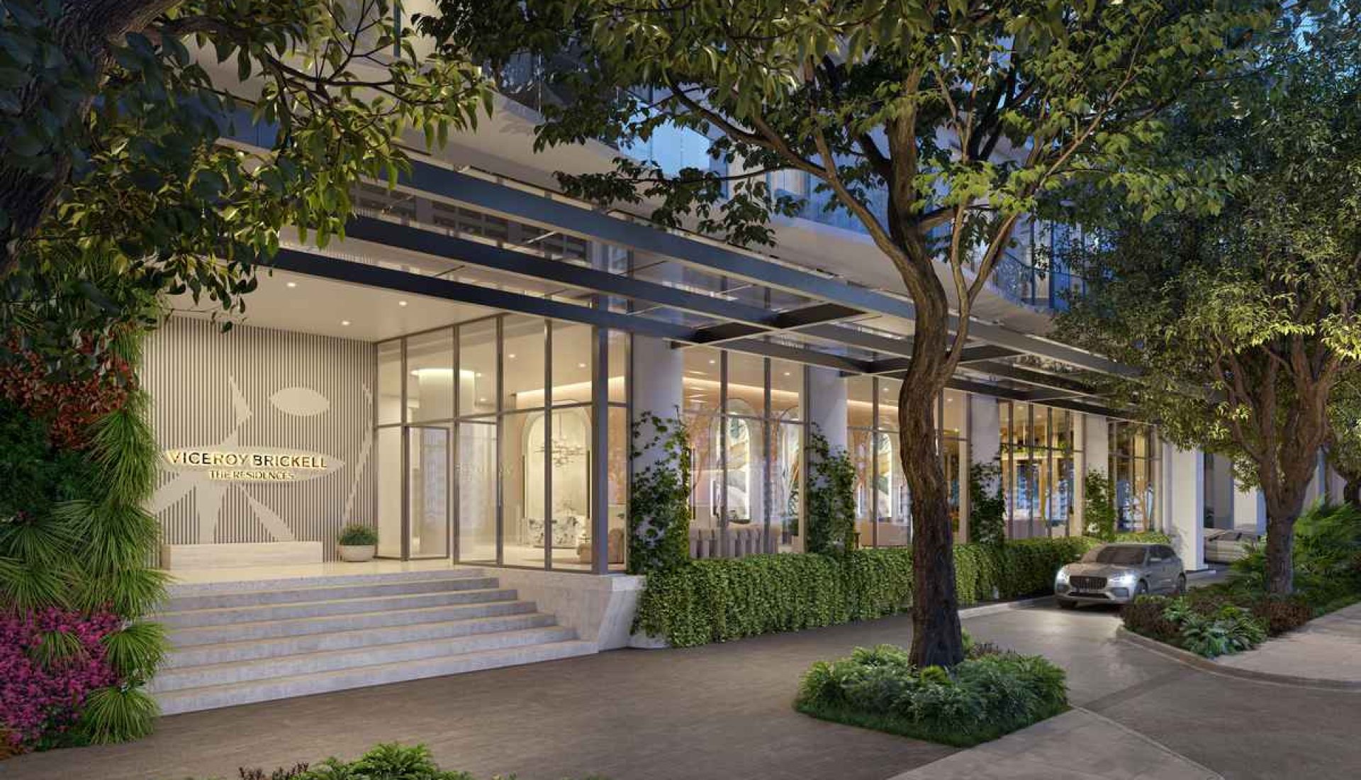 Entrance to a modern luxury building with glass walls, steps, and surrounding greenery including trees and shrubs.