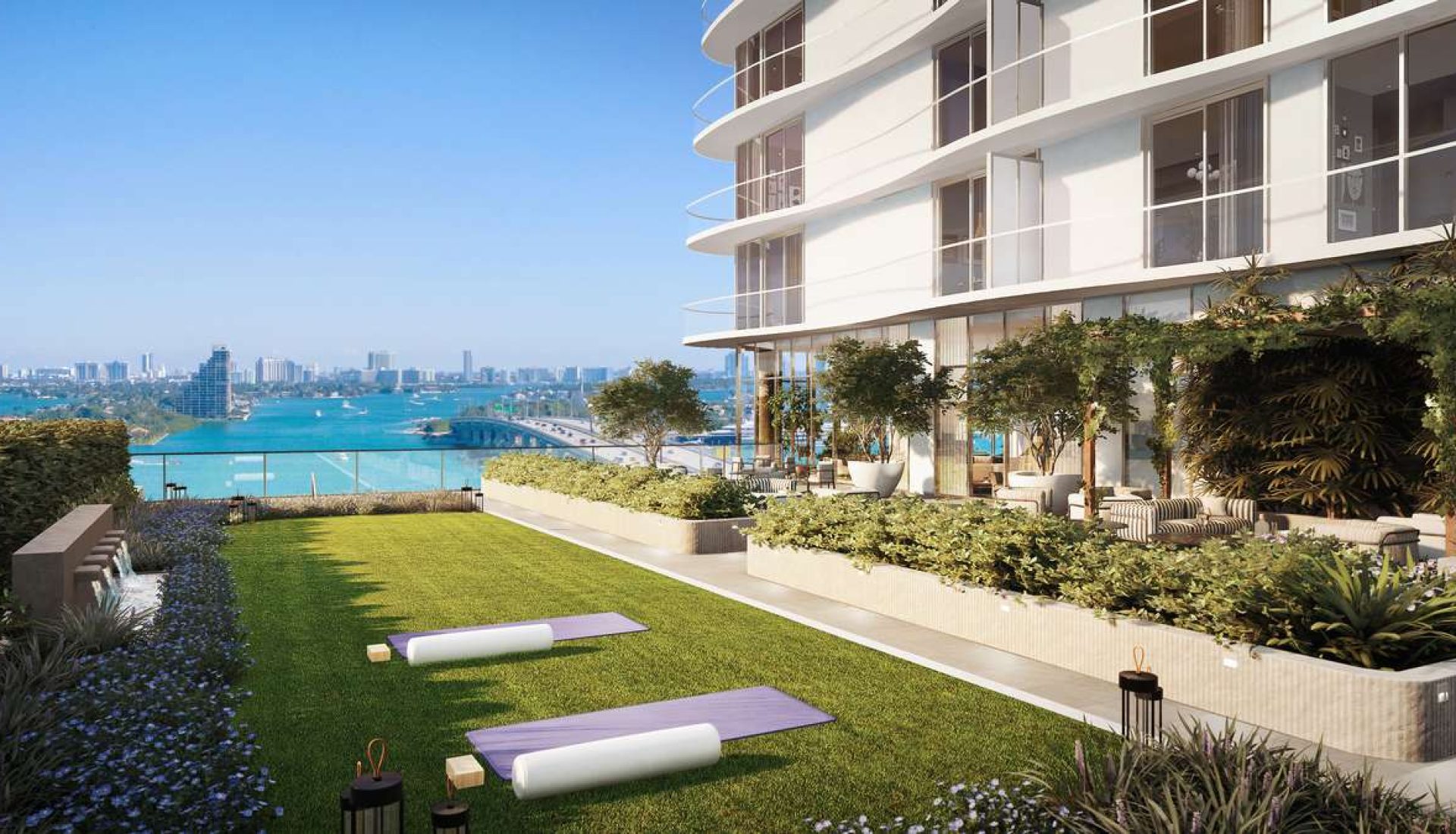 Rooftop garden of a modern white building with yoga mats on a green lawn and water views beyond a glass railing.