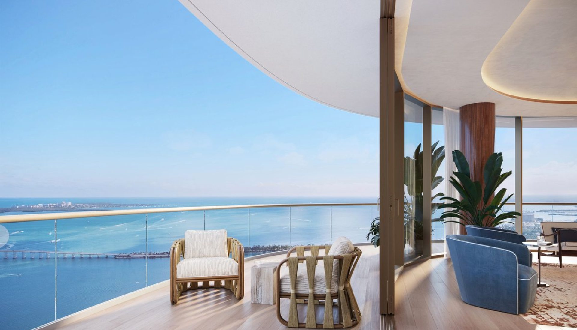 Sunlit balcony with glass railing overlooking a blue ocean, wicker chairs, and a small stone table.