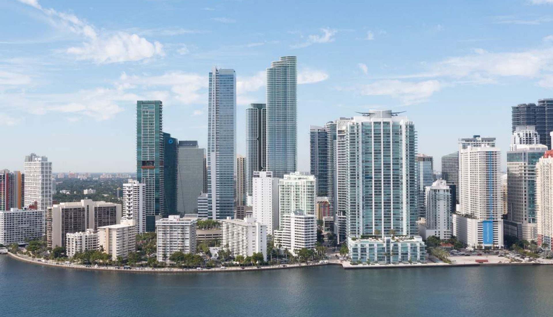 Skyline of modern high-rise buildings along a calm waterfront under a blue sky with scattered clouds, featuring glass towers and a curved shoreline, likely a coastal city center.