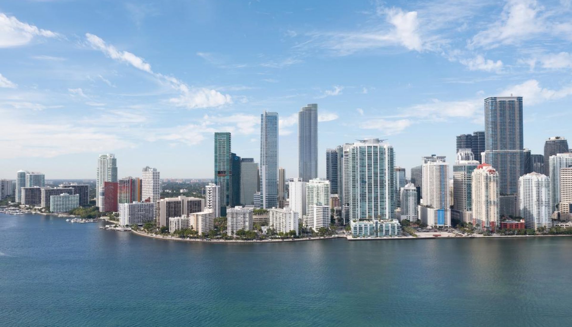 City skyline with tall glass skyscrapers along a calm blue harbor under a bright sky.