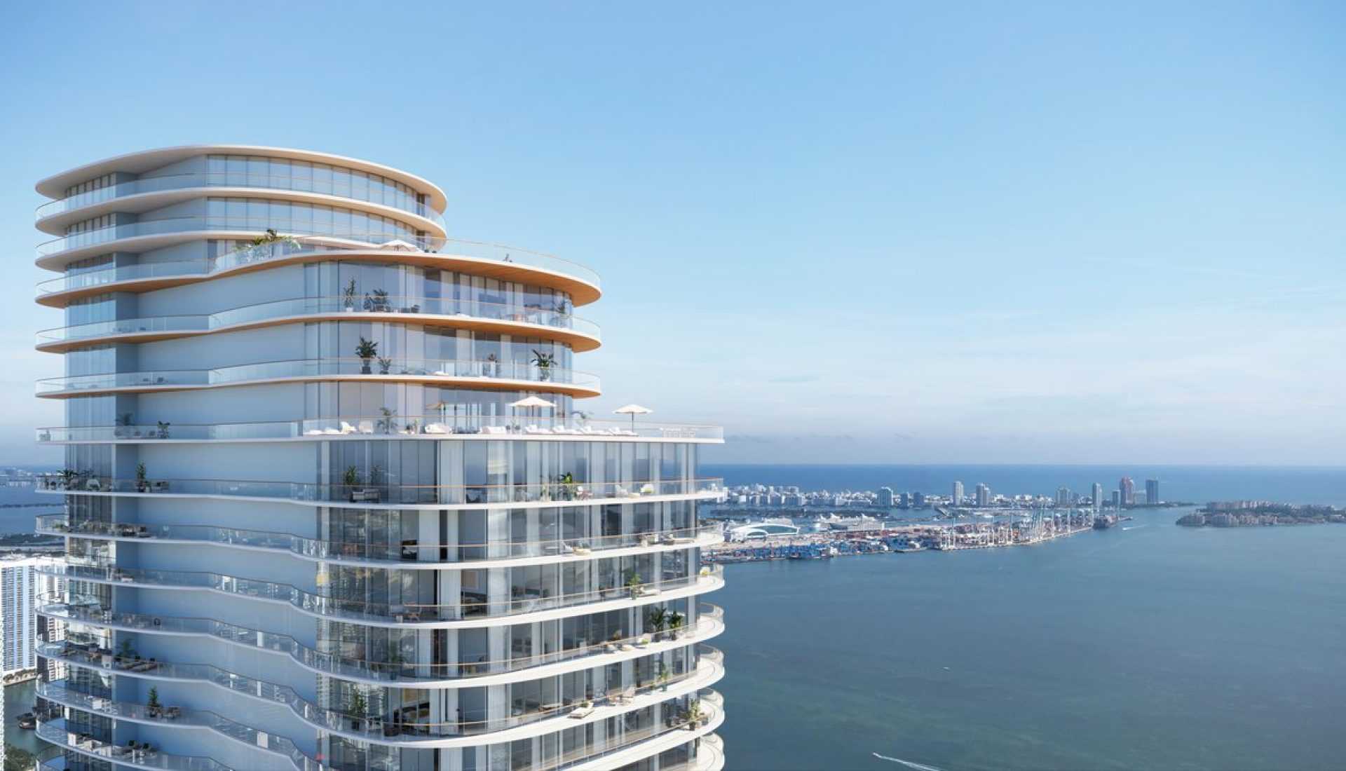 Curved glass residential tower with multiple terraces overlooking a harbor and distant city skyline.