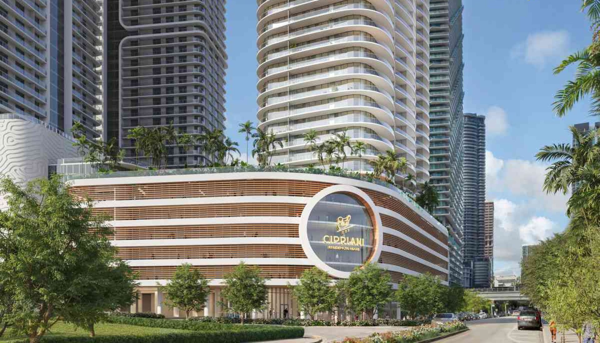 Urban streetscape with tall curved residential towers and a circular striped podium building with a logo, palm trees, and a landscaped median.